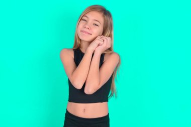 Yeşil arka planda siyah spor giysiler giyen sevimli sarışın küçük kız el ele tutuşup kameraya gülümsüyor.