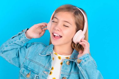 Mavi arka planda kot ceket giyen küçük sarışın kız gülümseyerek en sevdiği müziği kablosuz kulaklıkla dinlediği için çok mutlu..