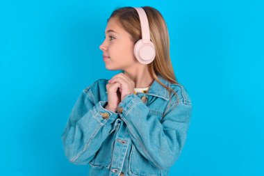 Sarışın, mavi arka planda kot ceket giyen küçük kız müzik kulaklığı takıyor. Konsantre olmuş müzik dinliyor ve ilgiyle yan tarafa bakıyor..