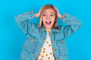 Mavi arka planda kot ceket giyen neşeli, neşeli sarışın küçük kız harika haberler aldıktan sonra ellerini havaya kaldırıyor..