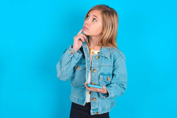 デニムジャケットを着用しているブロンドの慎重な少女は 何かについて深く考え 現代の携帯電話を使用して 良いメッセージを構成しようとし 唇の近くにインデックス指を保ちます — ストック写真