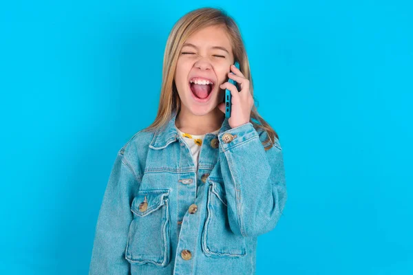 ジーンズジャケットブルーバックグラウンドを身に着けている幸せな小さな白人少女は 電話会話中に友人からの面白い話を積極的に聞きます — ストック写真