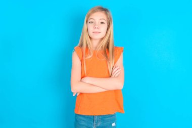 Kendine güveni tam, sakin, turuncu tişörtlü, mavi arka planda kolları bağlı bir kız çocuğu. Profesyonel titreşimlerin kendine güvenen pozlarda durduğunu gösterir.