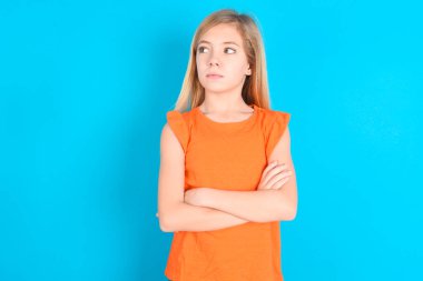 Mavi arka planda turuncu tişört giyen düşünceli küçük kız kollarını kavuşturmuş düşünceli bir yüz ifadesiyle ne yapacağını düşünüyor.