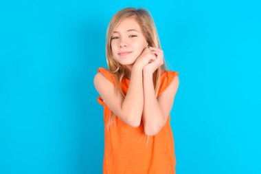 Mavi arka planda turuncu tişört giyen sevimli, ciddi bir kız çocuğu kameraya nazikçe gülümsüyor.