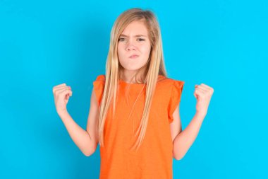 Mavi arka plan üzerine turuncu tişört giyen sinirli küçük kız yanakları öfkeyle emiyor ve yumruklarını kaldırıyor öfke ve agresif duyguları ifade ediyor. Öfkeli model