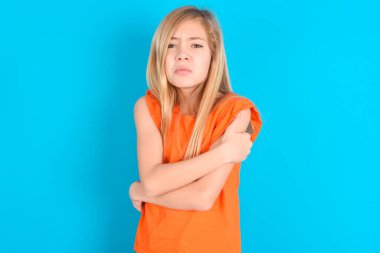 Mavi arka planda turuncu tişört giyen küçük bir kız. Kış soğuğu yüzünden titriyor ve donuyor. Yüzünde üzgün bir ifade var..