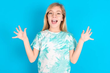 Mavi arka planda modaya uygun tişörtler giyen küçük beyaz kız harika bir sonuç elde ettikten sonra ağzını ve kollarını açar.