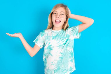 Şaşırmış küçük beyaz kız mavi arka planda moda tişörtler giyip el ele tutuşup teklif sunuyor.