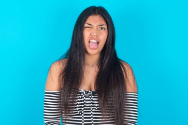 Vücut dili. Modaya uygun giyinen, mavi arka planda kaşlarını çatan, tiksindirici bir Latin kadın..