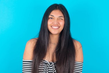 Mavi arka planda giyinmiş Latin bir kadın mutlu bir ifadeyle göz kırpıyor. Yüz ifadeleri ve insanların duygu konsepti.