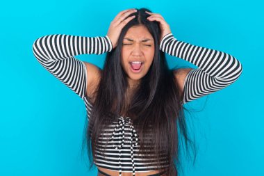 Şok olmuş, mavi arka planda giyinmiş, elleri başının üstünde umutsuzluk ve hüsran içinde bağıran Latin bir kadın..