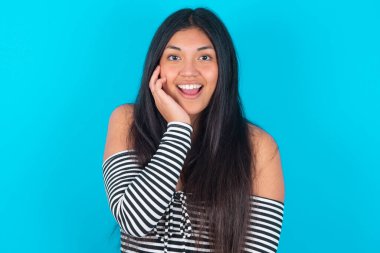 Şok olmuş, afallamış, mavi arka planda moda kıyafetleri giyen, ağzı açık, ağzı açık, eli yüzüne yakın Latin bir kadın. Olumlu duygu yüz ifadesi vücut dili.