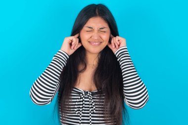 Bu sinir bozucu sesi çıkarmayı kes! Mutsuz, stresli, latin bir kadın, mavi arka planda moda kıyafetleri giyiyor, endişeli bir yüz ifadesi takınıyor, kulaklarını parmaklarıyla tıkıyor, gürültüden rahatsız oluyor..