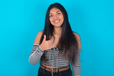 Mavi arka planda giyinmiş Latin bir kadın baş parmağını kaldırıyor, aferin! Olumlu insani duygu yüz ifadesi vücut dili.