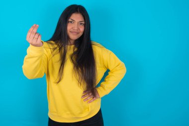 What the hell are you talking about. Shot of frustrated  latin woman wearing yellow sweatshirt over blue background gesturing with raised hand doing Italian gesture, frowning, being displeased and confused with dumb question.