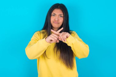 Mavi arka planda sarı eşofman giyen Latin bir kadın reddedilmiş kızgın bir ifadeyle negatif işaret veriyor..