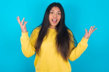 Neşeli, şanslı Latin kadın mavi arka planda sarı kazak giyiyor tezahürat yapıyor, başarıyı kutluyor, yumruklarını sıkarak evet diye bağırıyor.