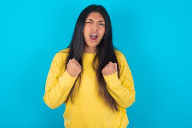 Neşeli, şanslı Latin kadın mavi arka planda sarı kazak giyiyor tezahürat yapıyor, başarıyı kutluyor, yumruklarını sıkarak evet diye bağırıyor.