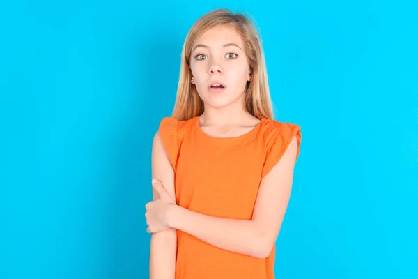 穿着橙色T恤蓝色背景的尴尬的小女孩张大了嘴 令人难以置信的新奇事物在呆呆地凝视着 — 图库照片