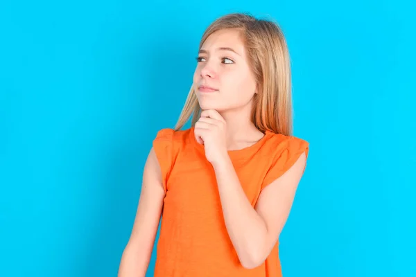 Bedachtzame Kleine Meisje Dragen Oranje Shirt Blauwe Achtergrond Houdt Kin — Stockfoto