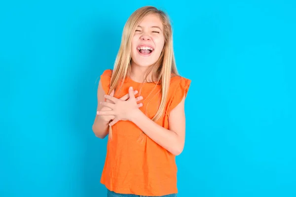 青い背景にオレンジ色のTシャツを着ている小さな子供の少女は幸せを表現し 喜んで笑い 心に手を置きます — ストック写真