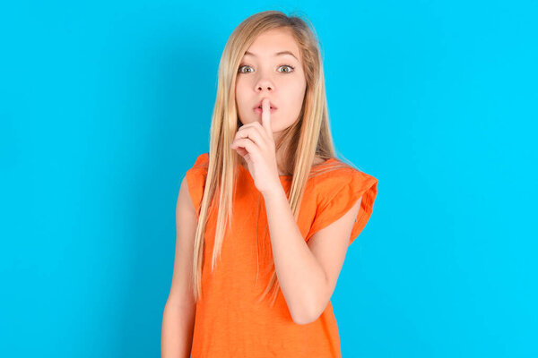 Удивлённая маленькая девочка в оранжевой футболке на синем фоне делает жест молчания, держит палец над губами и смотрит таинственно в камеру