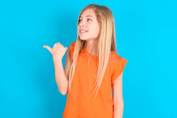 Liten Flicka Bär Orange Shirt Över Blå Bakgrund Pekar Bort — Stockfoto