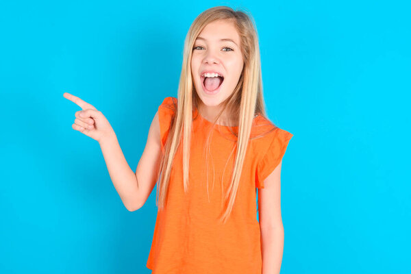 little kid girl wearing orange T-shirt over blue background points aside on copy blank space. People promotion and advertising concept