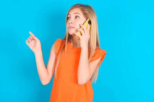 Маленькая Девочка Оранжевой Футболке Синем Фоне Говорит Мобильному Телефону Проводит — стоковое фото