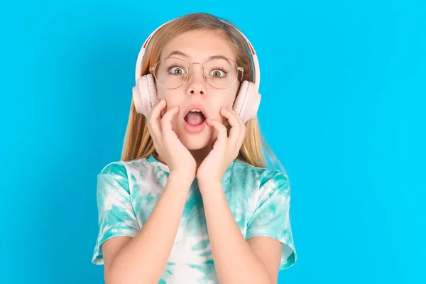 衝撃を受けた小さな慎重な少女はカメラで怖がり 口を広く開け 耳にワイヤレスステレオヘッドフォンを着用する — ストック写真