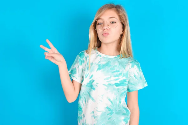 little caucasian kid girl wearing trendy T-shirt over blue background makes peace gesture keeps lips folded shows v sign. Body language concept