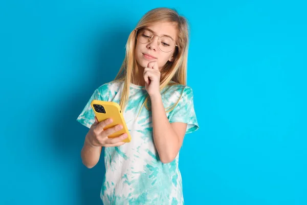 青い背景にトレンディなTシャツを着ている思慮深い幸せな小さな白人少女は 携帯電話 コピースペースを見ます — ストック写真