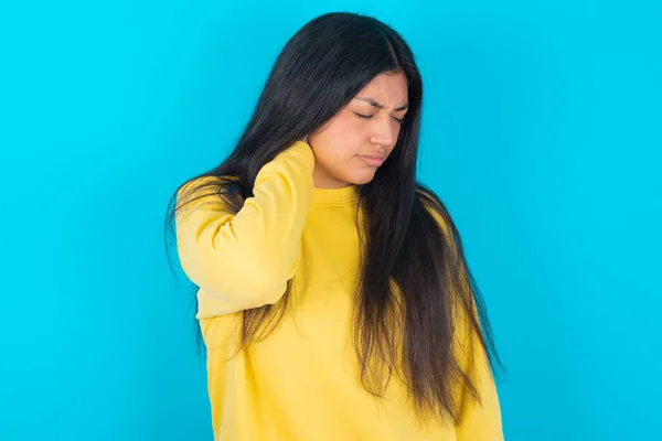 latin woman wearing yellow sweatshirt over blue background suffering from back and neck ache injury, touching neck with hand, muscular pain.