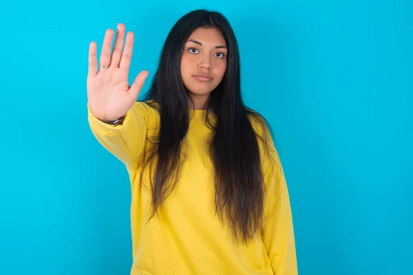 latin woman wearing yellow sweatshirt over blue background doing stop gesture with palm of the hand. Warning expression with negative and serious gesture on the face.