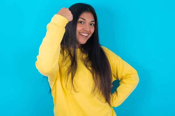 青色の背景に黄色いスウェットシャツを着ているオーバージョイスラテン女性は 良いニュースを受け取って喜んで ひび割れ 勝利のジェスチャーを作ります — ストック写真