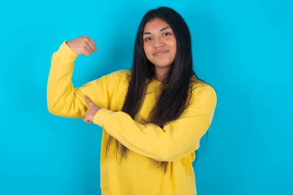 身披黄色运动衫 身披蓝色背景 面带微笑的拉丁女子举手表决肌肉 自信胜利 坚强而独立 — 图库照片