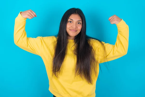 蓝色背景下穿着黄色运动衫的拉丁女子抬起胳膊 展示了她对胜利的信心 看上去坚强而独立 对着相机笑得积极 体育概念 — 图库照片