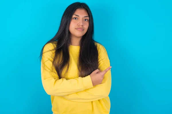 latin woman wearing yellow sweatshirt over blue background smiling broadly at camera, pointing fingers away, showing something interesting and exciting.