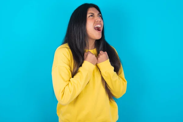latin woman wearing yellow sweatshirt over blue background excited and glad to achieve victory, clenches fists, screams in excitement with closed eyes,successful person.