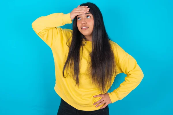 latin woman wearing yellow sweatshirt over blue background touching forehead, hears something surprising, glad receive good news, feels relieved. Almost got in trouble.