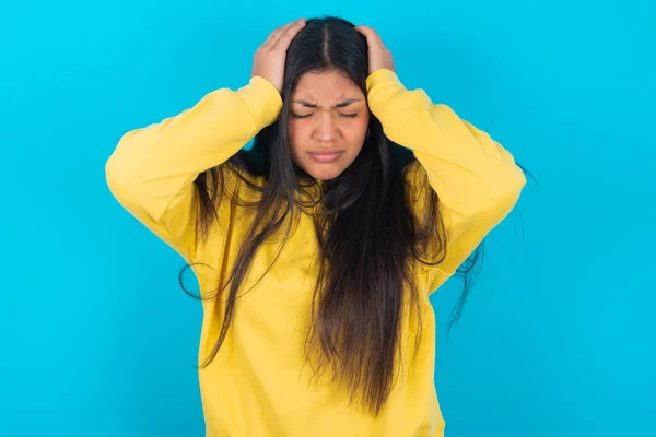 latin woman wearing yellow sweatshirt over blue background holding head with hands, suffering from severe headache, pressing fingers to temples