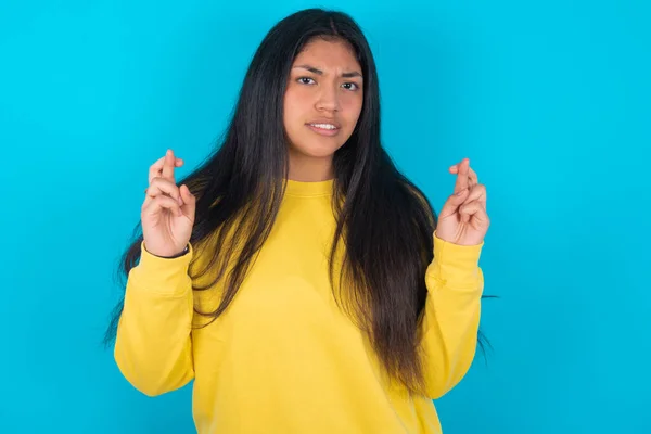 latin woman wearing yellow sweatshirt over blue background holding fingers crossed with worried expression hoping boss didn\'t noticed mistakes at work.