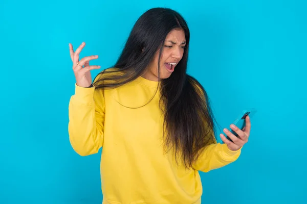 Photo of outraged annoyed latin woman wearing yellow sweatshirt over blue background holds cell phone, makes call, argues with colleague,  expresses negative emotions. People and anger.