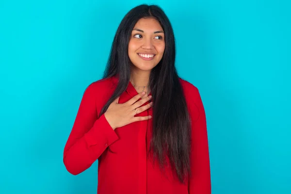 青色の背景に赤いTシャツを着ている楽しいラテン語の女性は 面白いことを思い出し 胸に手を伸ばし 幸せに笑います — ストック写真