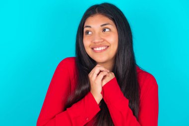 Mavi arka planda kırmızı tişört giyen mutlu Latin kadın harika bir şey olacağını tahmin ediyor, mutlu bir şekilde yan yana bakıyor, ellerini yan yana tutuyor, mutlu bir ifadeye sahip..