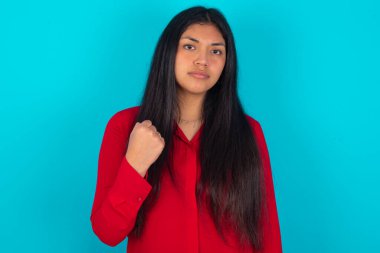  Mavi arka planda kırmızı tişört giyen Latin bir kadın yumruğunun, intikam almak ya da birini tehdit etmek için sinirli bir yüz ifadesi olduğunu gösteriyor. Sana patronun kim olduğunu göstereceğim.