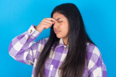 Mavi arka planda ekose gömlek giyen üzgün Latin kadın baş ağrısından muzdarip.