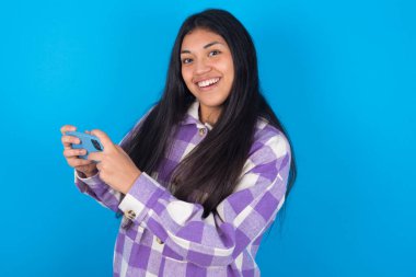 İyi bağımlı, neşeli Latin kadın. Mavi arka planda ekose gömlek giyiyor. Ağ oyunu oynuyor.