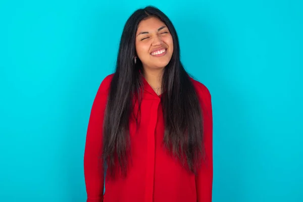 青い背景に赤いTシャツを着たポジティブなラテン語の女性は 大喜びの表情で目を閉じ 笑いは白い完璧な歯を示しています — ストック写真